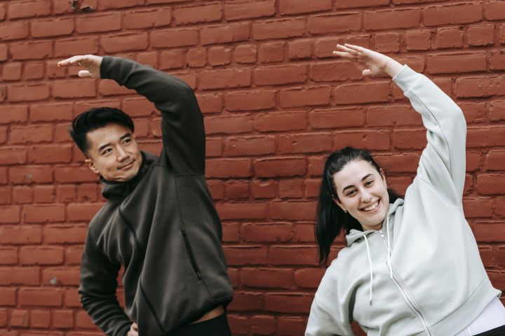 Positive young obese woman and Asian male trainer in sportswear warming up before outdoor training standing near brick wall